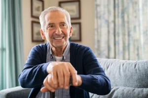 An elderly man with a warm smile sits on a couch, resting his hands on a cane. The well-lit room features framed pictures and patterned curtains, creating a cozy atmosphere.
