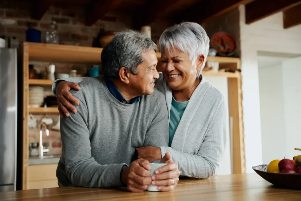 An elderly couple shares a warm and joyful moment in a cozy kitchen. The man, wearing a gray sweater, holds a cup while his partner, dressed in a light cardigan, embraces him affectionately. They look at each other with love and happiness, surrounded by a homely atmosphere with wooden shelves, kitchenware, and a bowl of fruit on the table.