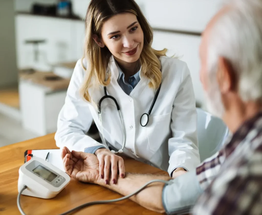 Smiling female doctor in a white coat checking an elderly patient's blood pressure with a digital monitor in a clinical setting.