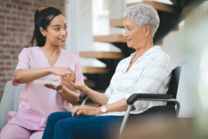 Smiling caregiver in pink scrubs assisting an elderly woman in a wheelchair by handing her medication in a well-lit home setting.