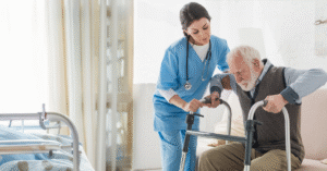A clinician in a skilled nursing facility helps an elderly male patient transitioning to the facility stand using a walker.