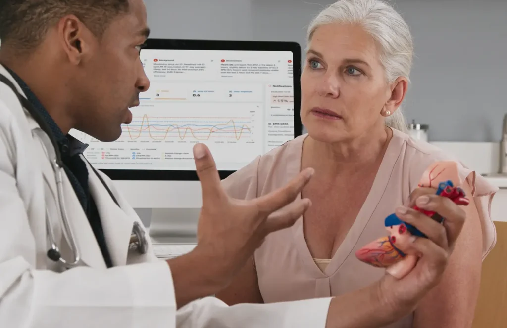 Cardiologist holds an anatomical presentation of the heart as he talks with his female patient in-office.