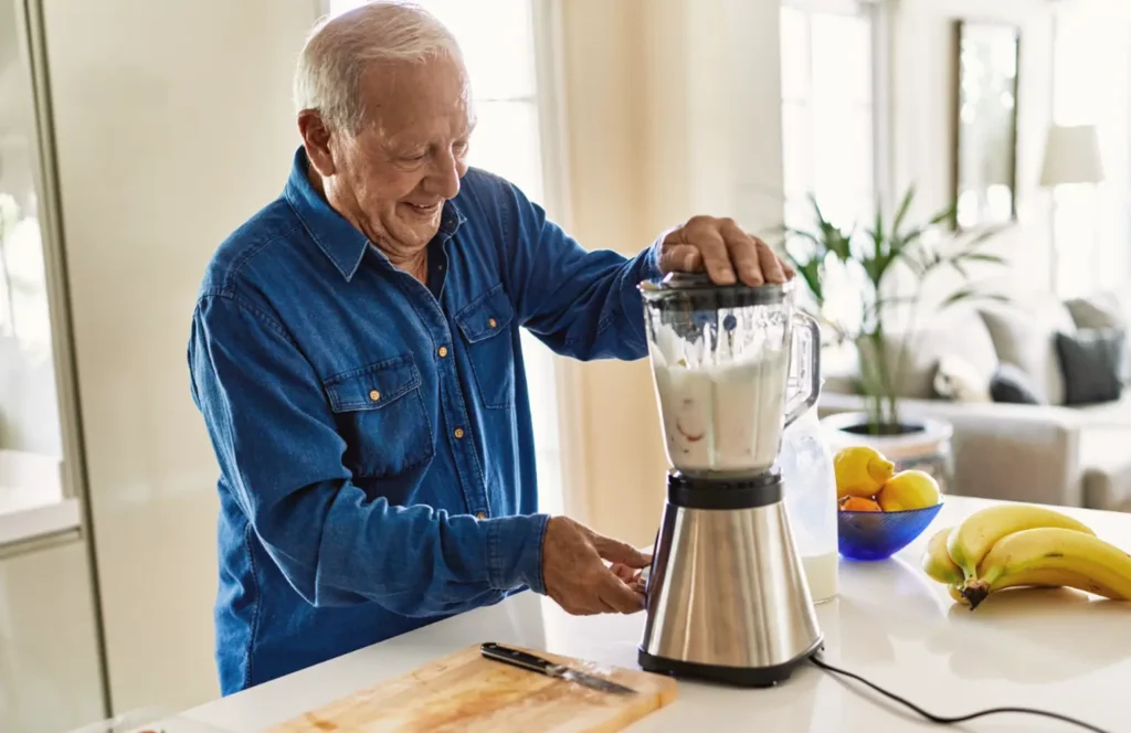 Elderly man uses a blender to make a fruit smoothie in the kitchen of his apartment in an independent living community.